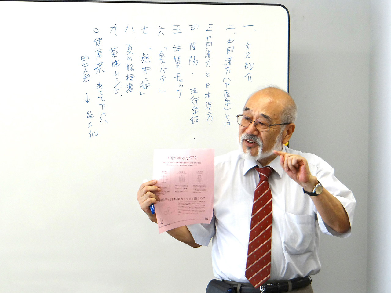 ホワイトボードを背に、片手にチラシを持って説明している種田生司。
